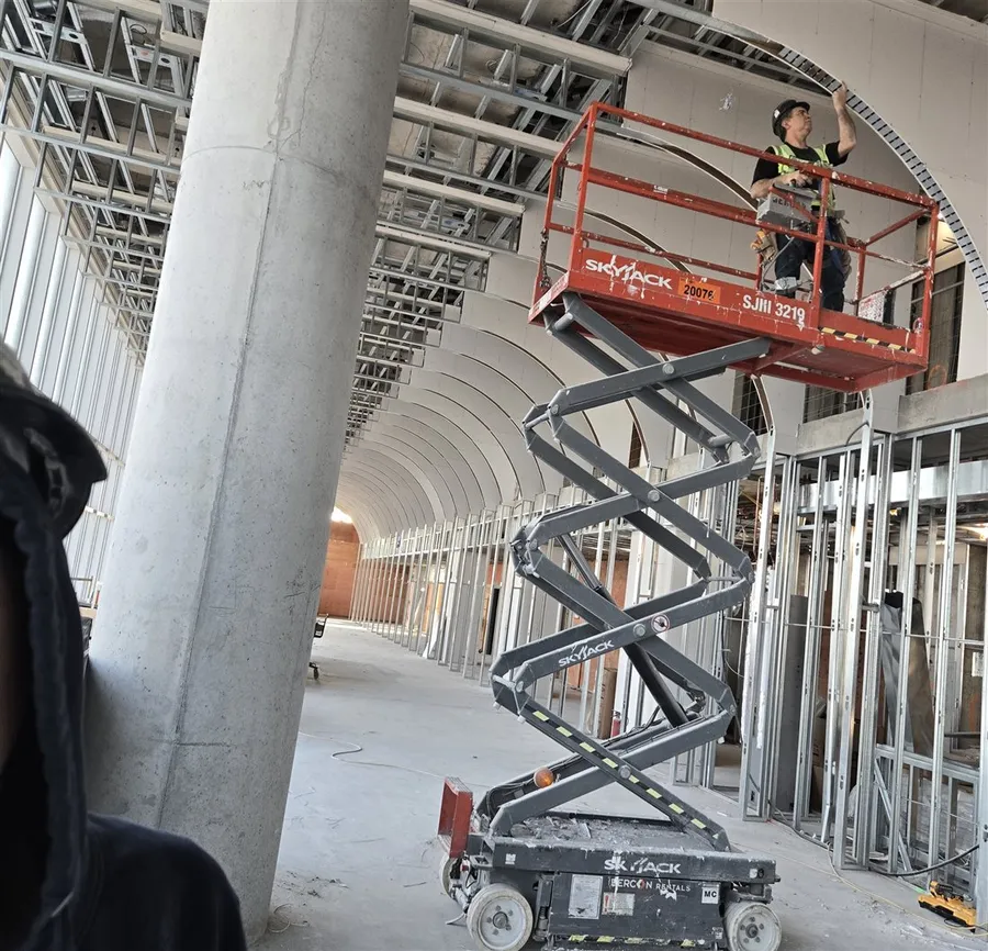 A scissor lift beside a concrete column under curved metal framing inside an unfinished commercial building