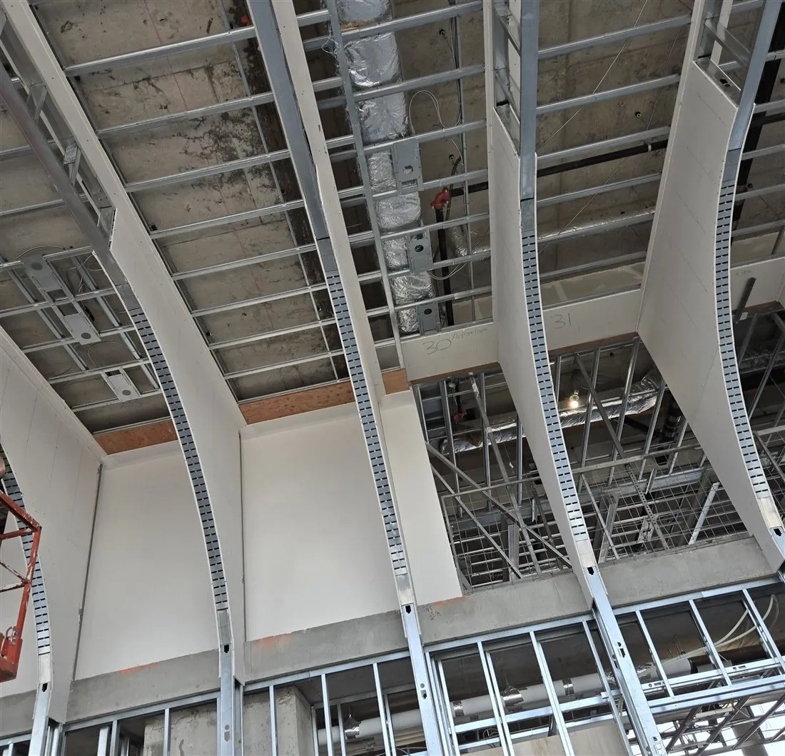 Curved metal framing and plasterboard along a long commercial floor with a concrete column and a scissor lift, photographed in May 2025.
