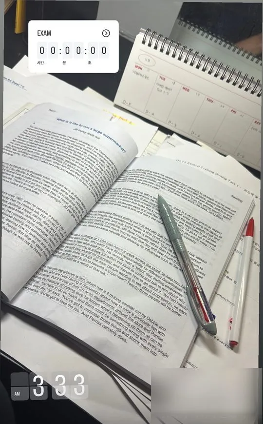A desk late at night with an open IELTS practice book, printed writing practice sheets, two pens and a weekly planner counting down to the exam. A clock reads 3:33 AM and an exam countdown reads zero.