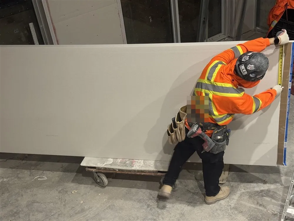 A drywall installer in a hard hat and hi-vis jacket, seen from behind, holding a tape measure against a full sheet of plasterboard standing on a trolley.