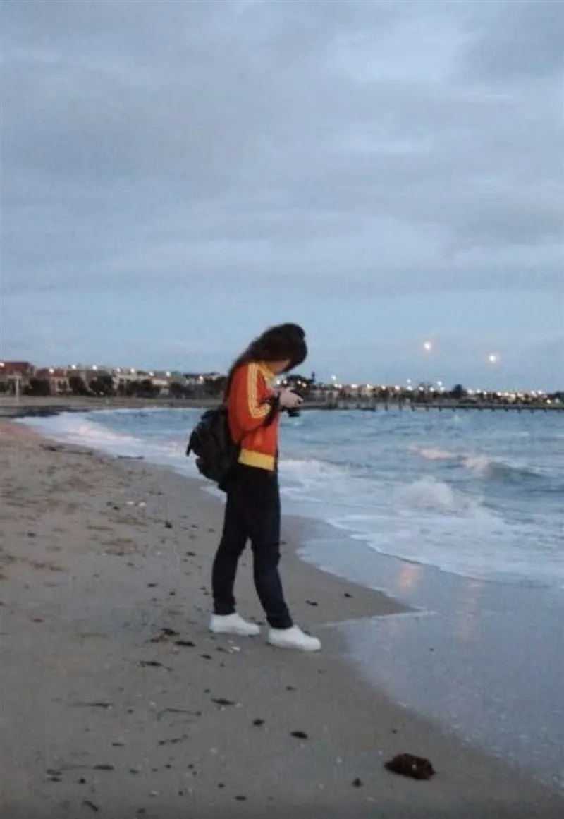 A person in an orange jacket with a backpack stands on St Kilda Beach at dusk, looking down at a camera, with the lights of the shoreline behind.