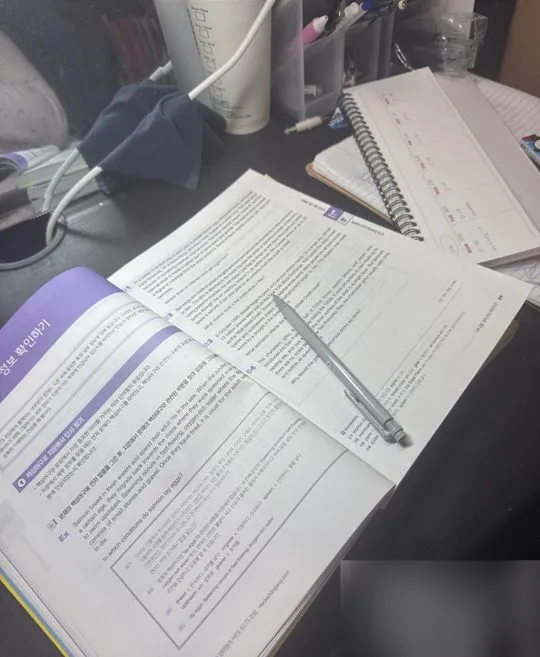 A study desk at night with an open English reading workbook, a pen, a face mask, a coffee cup and a weekly planner.
