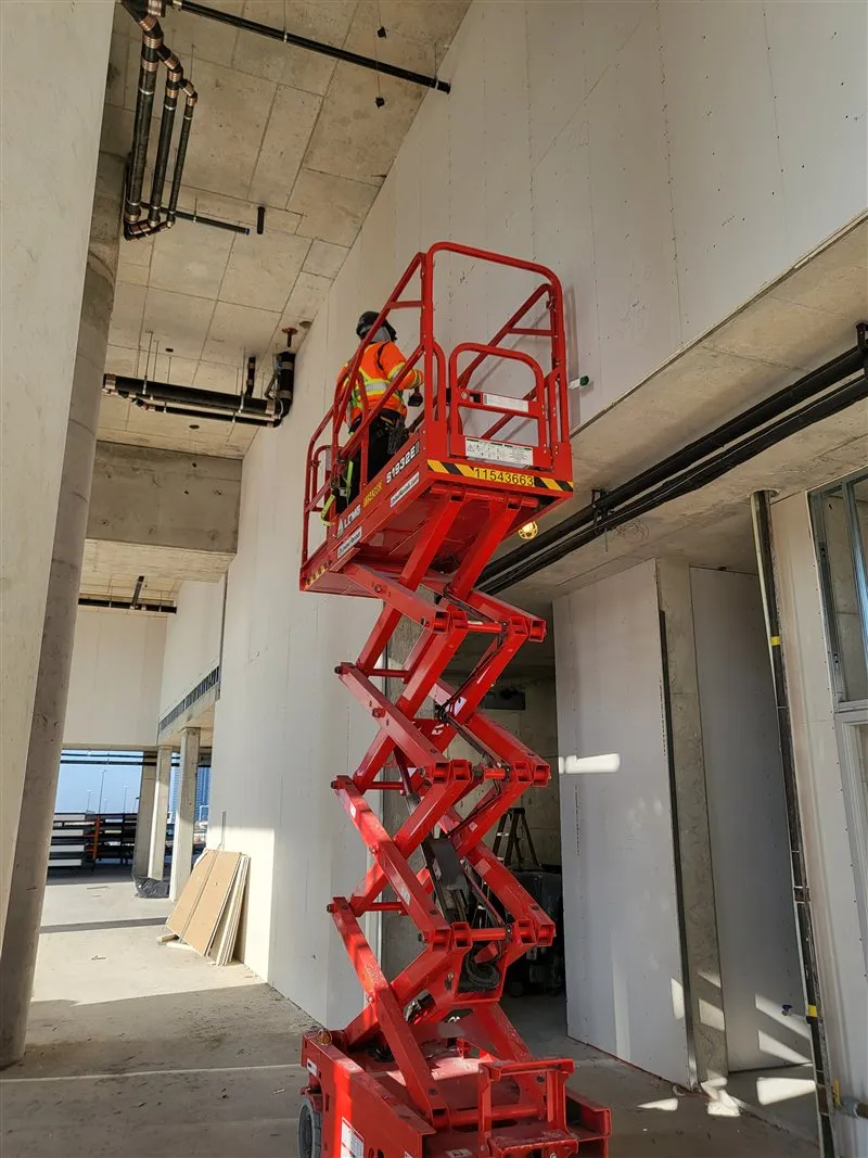 A worker in hi-vis on a red scissor lift beside a tall plasterboard wall on a commercial site, photographed in November 2023.